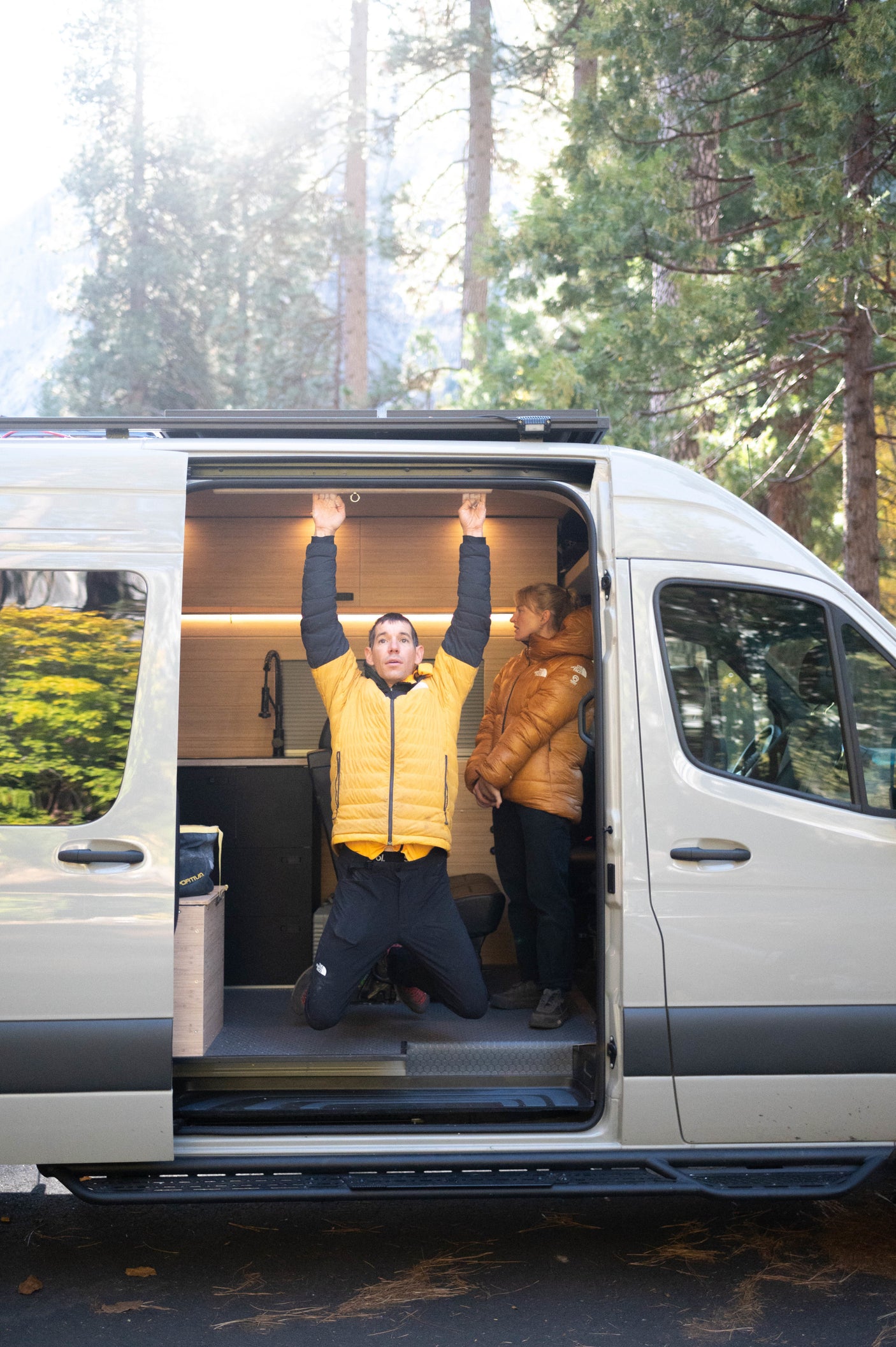A man trains, hanging from an RV rental camper van doorframe, as a woman watches nearby. Outdoor setting.