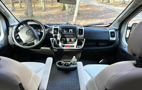 Spacious RV rental interior featuring the dashboard, steering wheel, and front seats, overlooking a forest road.