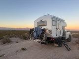 Overland RV rental: white truck camper with bikes and steps, parked on a gravel path in a desert landscape at sunset.