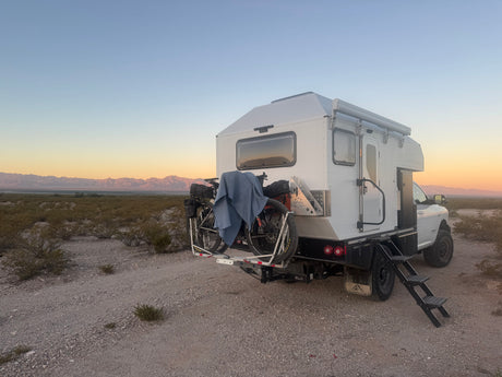 Overland RV rental: white truck camper with bikes and steps, parked on a gravel path in a desert landscape at sunset.