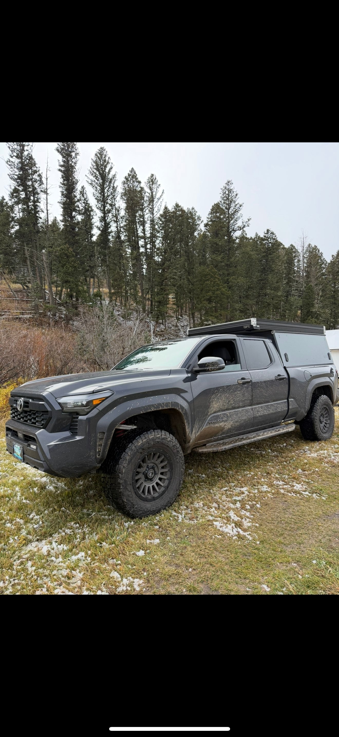 Dark grey Tacoma with a pop-top camper, muddy from trails, parked in a snowy forest. Ideal for an adventure RV rental.