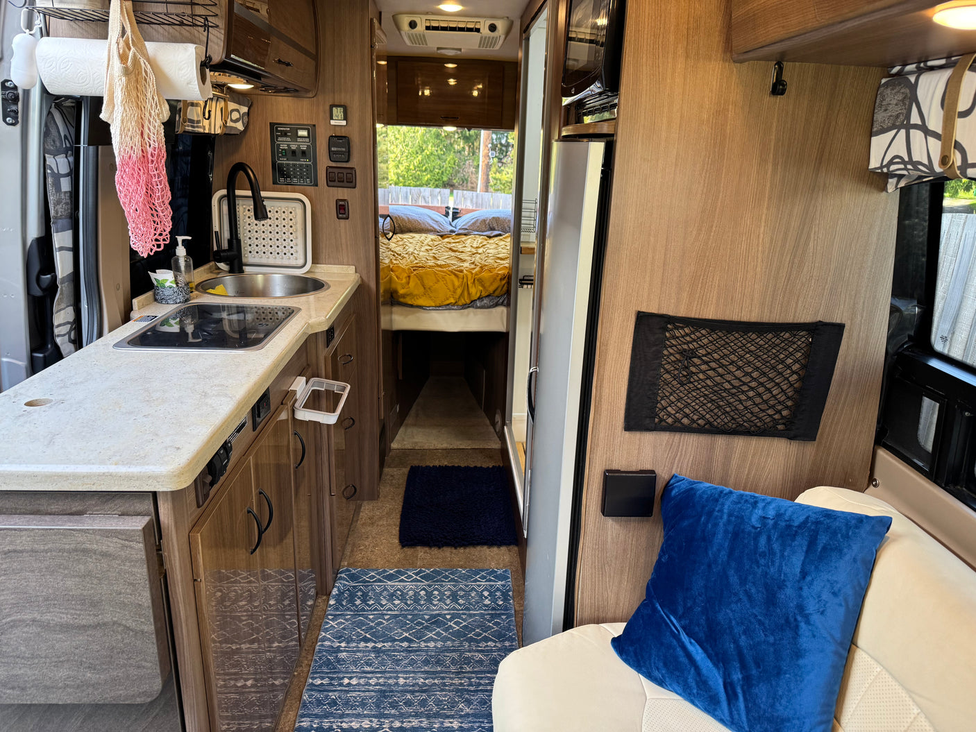 View down an rv rental interior: compact kitchen, cream seating with blue pillow, and bed in the back. Ideal for travel.