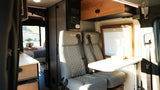 Inside a modern RV rental: gray Revel seats, dining table, and rear bedroom. Control panels and storage also visible.