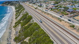 Aerial view of an RV rental on a coastal road next to train tracks, with ocean, beach, and homes along the bluff.