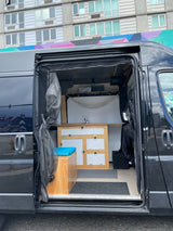 RV rental: Inside this black camper van, discover its compact kitchen, wooden storage, and a bench with a blue cushion, ideal for mobile living.