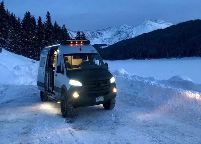  overlooking ski slopes and a town.|White Winnebago Revel RV rental parked on a sandy beach by the ocean. A dog sits inside the driver's seat