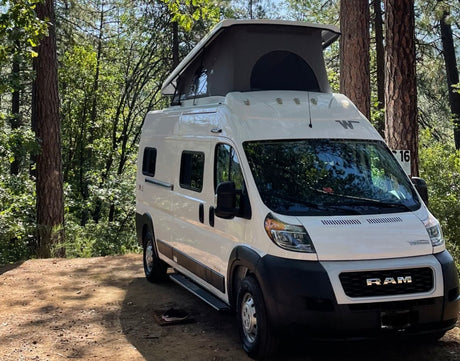 White camper van (RV rental) with a pop-top roof, parked at campsite #16 in a sunny, wooded area with tall trees. Ideal for nature camping.