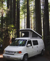White pop-top camper van, an RV rental option, parked under towering forest trees for outdoor adventure.