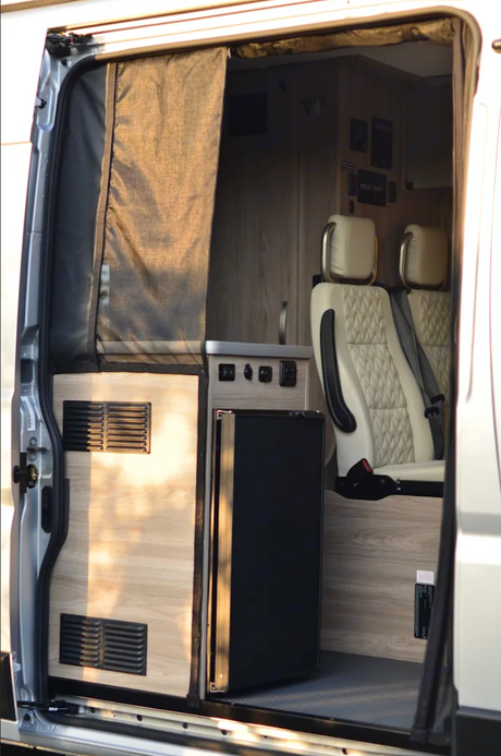 This RV rental's inviting interior reveals a compact kitchen with fridge, stylish passenger seating, and a convenient privacy curtain.