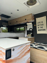 RV rental interior: modern, dark walls, light wood cabinets. Features a comfy bed, compact kitchen, and mountain decor.