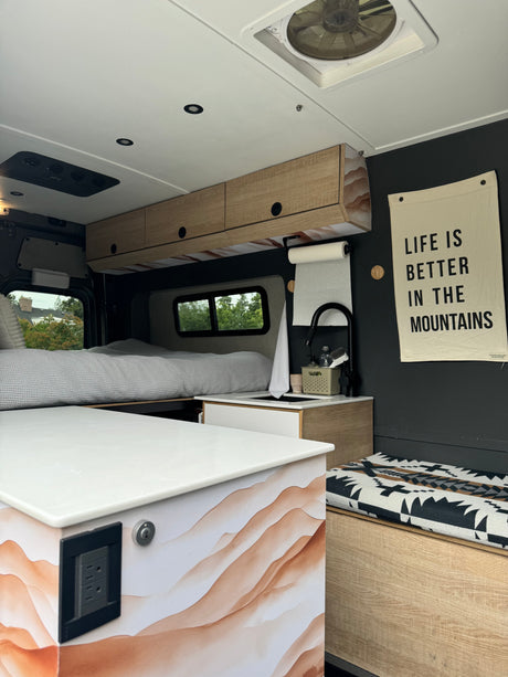 RV rental interior: modern, dark walls, light wood cabinets. Features a comfy bed, compact kitchen, and mountain decor.