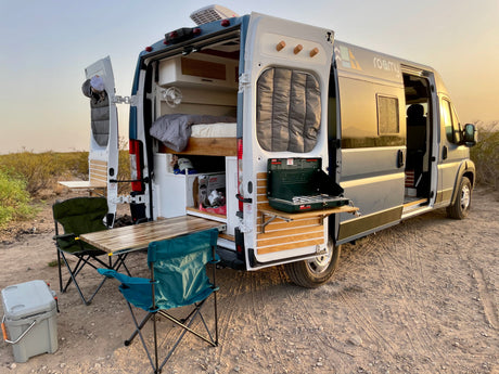 Roamy RV rental van with open back, revealing bed, outdoor kitchen, table, and chairs for camping.