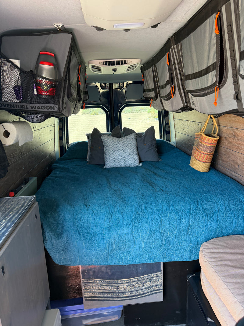 Interior of an **rv rental** featuring a cozy bed with a blue quilt, overhead storage, and rear windows with an outdoor view. — photo 16