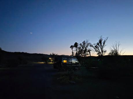 RV rental at twilight. A camper van with glowing windows is parked at a dark campsite, silhouetted by trees & hills.