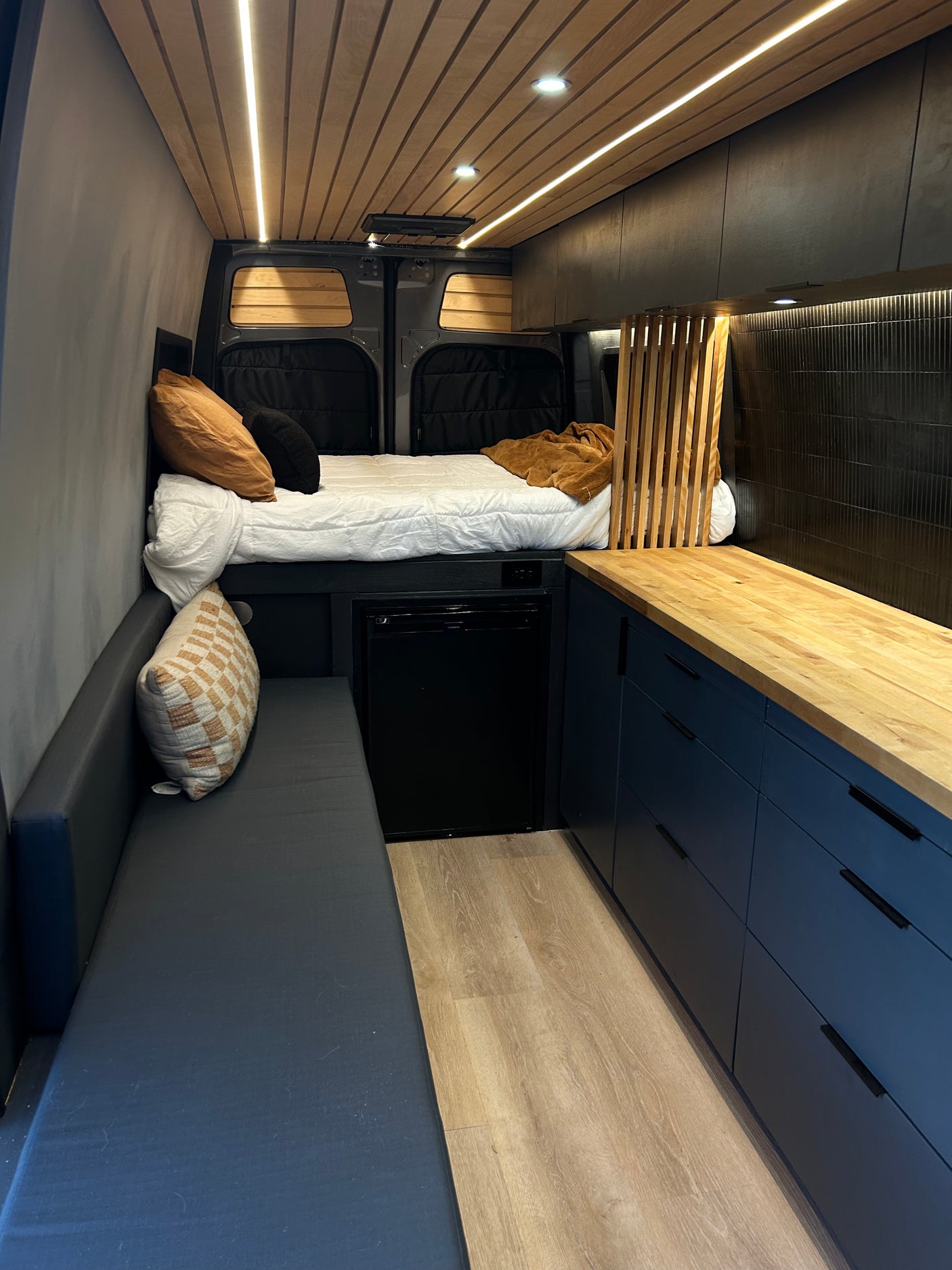 Modern RV rental interior with a cozy bed, sleek kitchen, and comfy lounge area, featuring wood ceilings and stylish finishes.