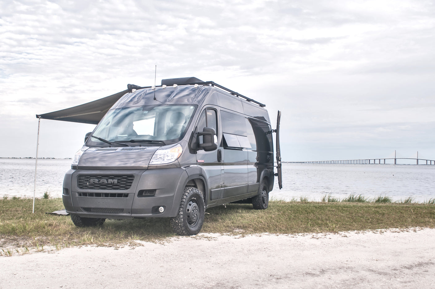 Explore scenic views! Dark grey Ram ProMaster RV rental van with awning extended, parked by water with a long bridge in the distance.