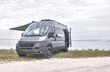 Explore scenic views! Dark grey Ram ProMaster RV rental van with awning extended, parked by water with a long bridge in the distance.