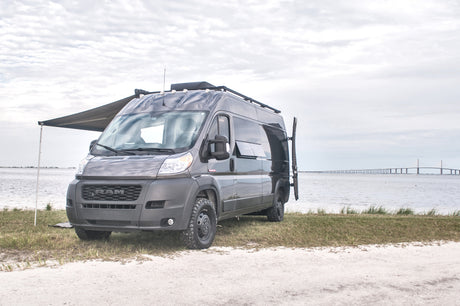 Explore scenic views! Dark grey Ram ProMaster RV rental van with awning extended, parked by water with a long bridge in the distance.