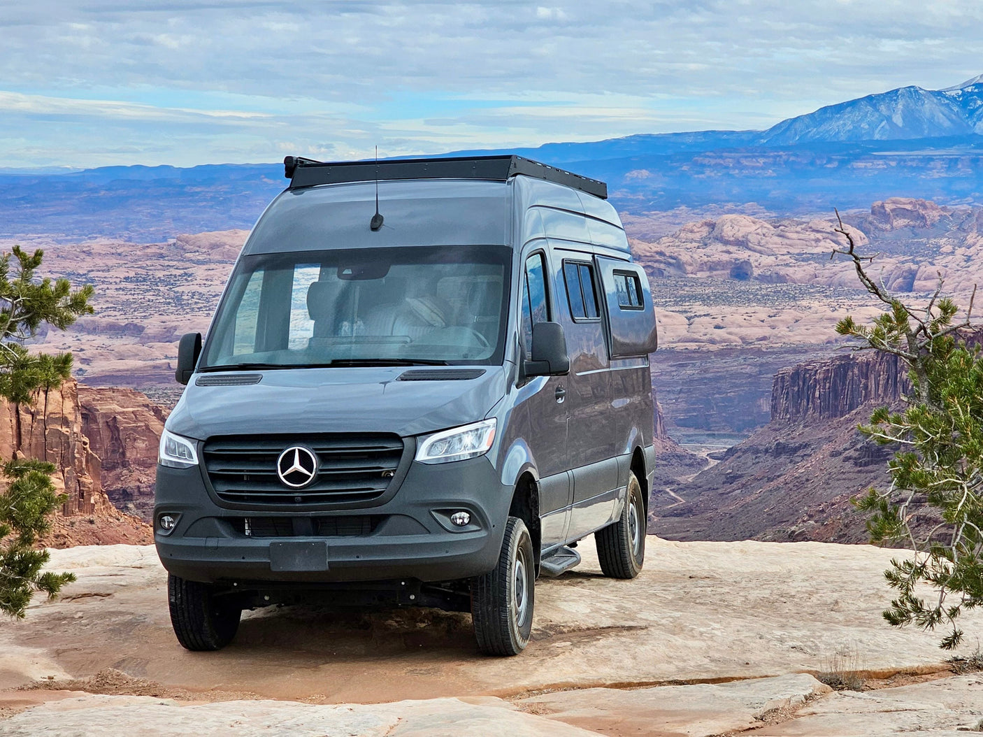 Gray Mercedes Sprinter RV rental camper van on a rocky ledge, overlooking vast desert canyons and distant mountains.