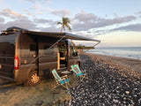 RV rental parked beachside at sunset. Dark camper van, awning out, two chairs on grass next to a pebbled beach with ocean & palm tree.