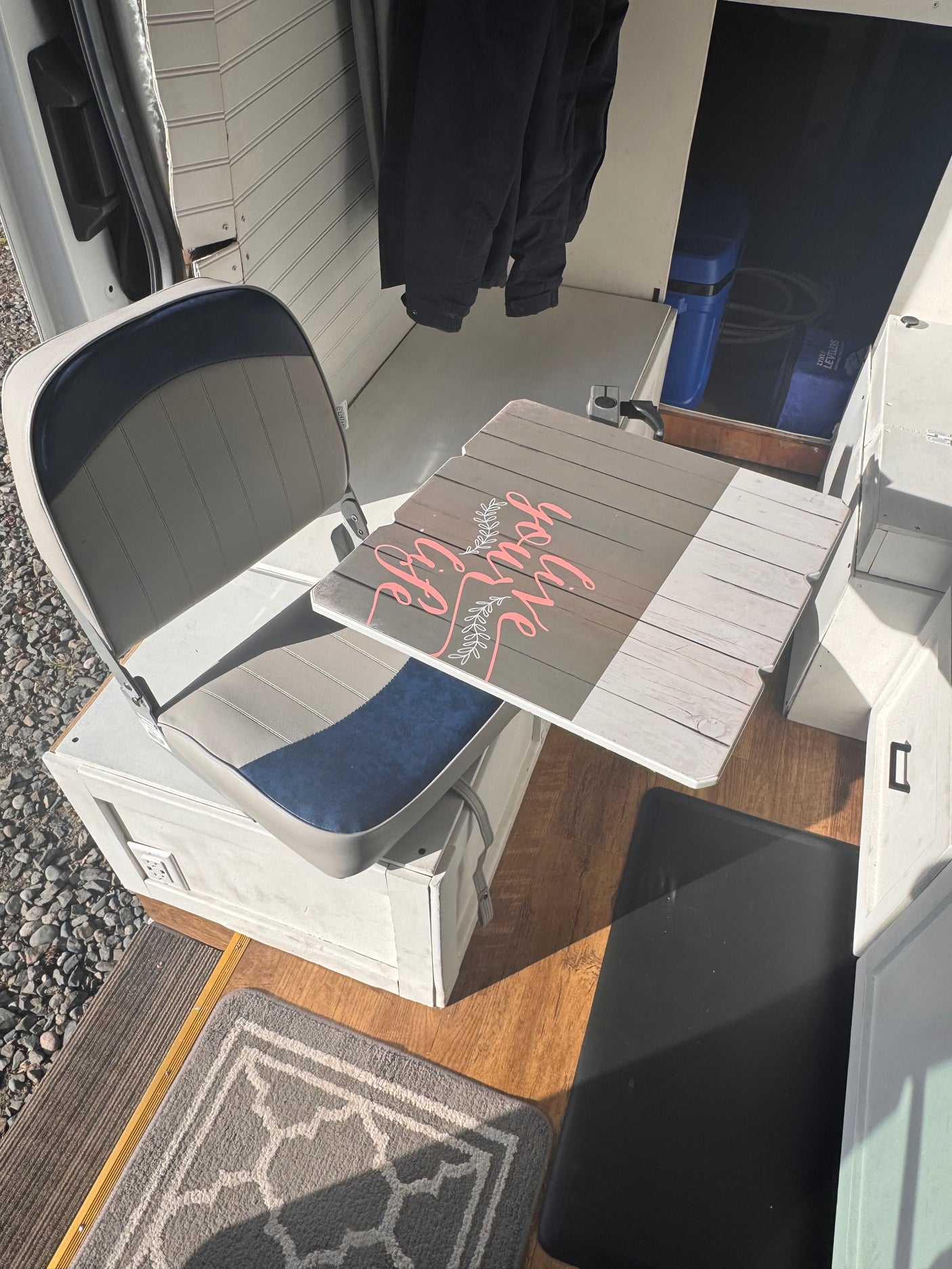 An RV rental's flexible interior: gray seat, wooden table with 'Live your life' art. Cozy travel setup.