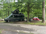 Dark grey camper van (RV rental) with rooftop tent at a wooded campsite, next to a ground tent and bikes.