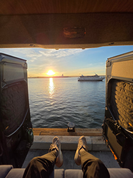 An RV rental view: feet up, enjoying golden hour sunset over the water from a campervan, with a boat.