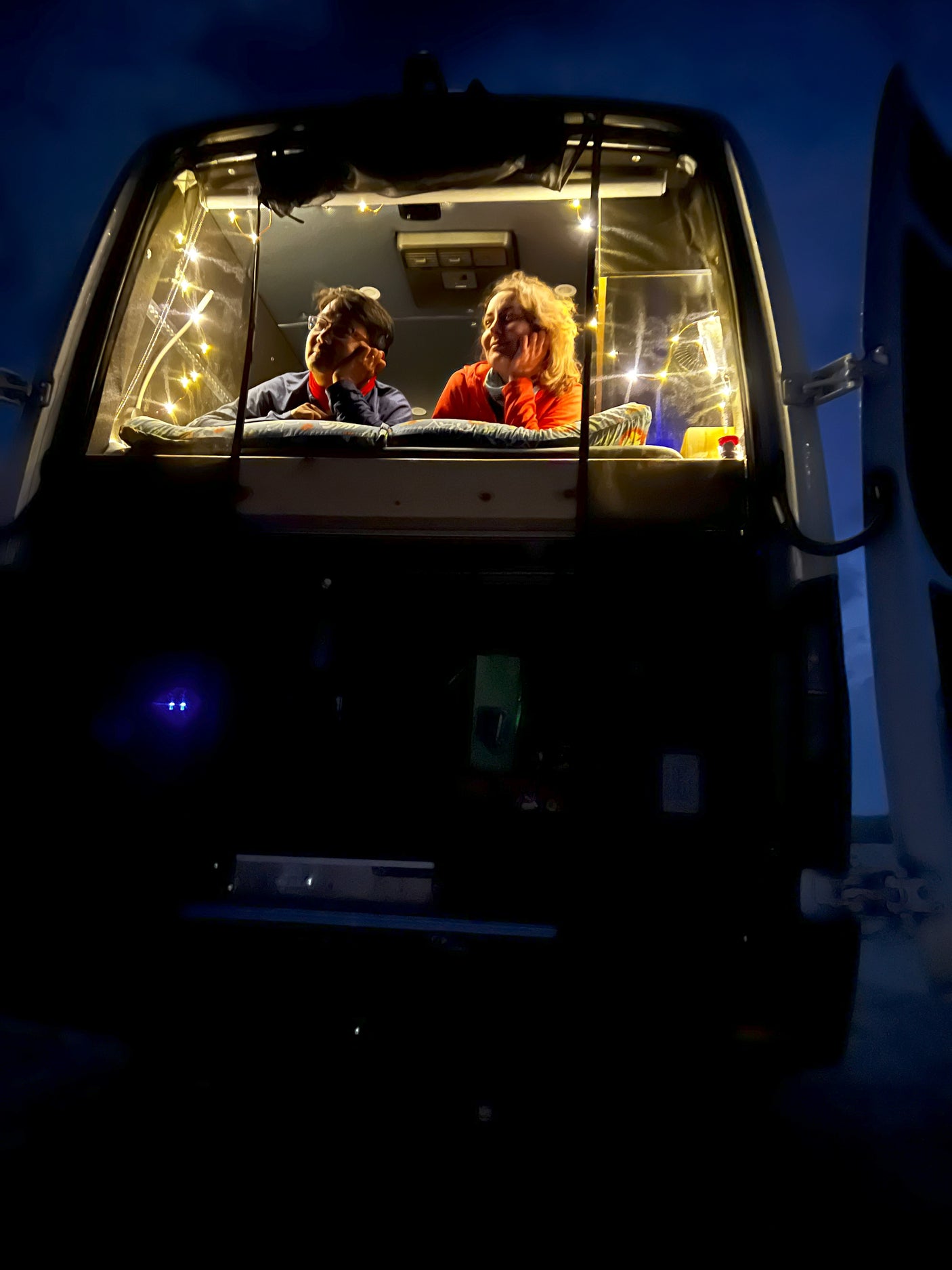Cozy night in an RV rental! Two people relax inside a camper van, illuminated by warm string lights, enjoying the view.