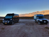 Two campers, an RV rental van & truck camper, parked in a desert landscape with mountains at sunset/sunrise.