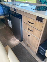 RV rental kitchen: light countertop, built-in stove/sink, microwave, compact fridge, & wood-look drawers.