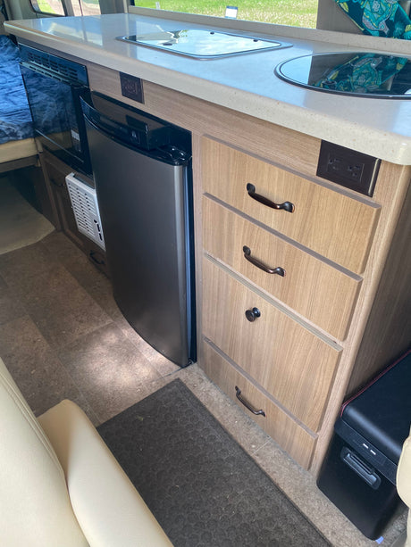 RV rental kitchen: light countertop, built-in stove/sink, microwave, compact fridge, & wood-look drawers.