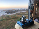 Kettle on stove in an RV rental van, facing a rocky coast. Perfect morning for camping & van life by the ocean.