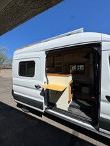 A white custom rv rental van with its sliding door open, revealing a cozy interior with a bed, shelves, and pull-out table.