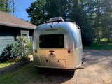 Airstream Bambi travel trailer, often available for rv rental, parked by a house amidst green trees.