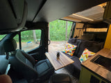 Spacious RV rental interior featuring swiveling front seats, a dining table, cozy lounge, and compact kitchen, with a forest view.