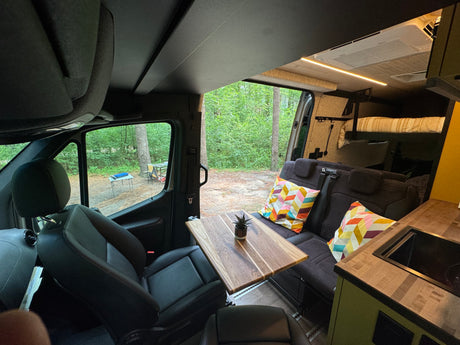 Spacious RV rental interior featuring swiveling front seats, a dining table, cozy lounge, and compact kitchen, with a forest view.