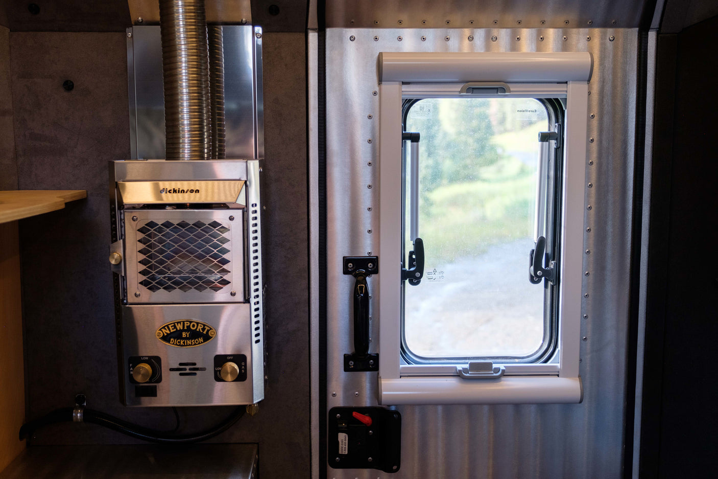 RV rental interior: Stainless steel Dickinson Newport heater & corrugated metal wall beside a window with a pull-down blind.