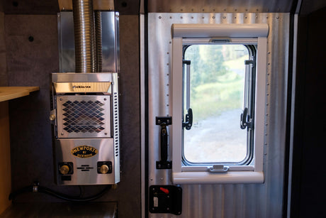 RV rental interior: Stainless steel Dickinson Newport heater & corrugated metal wall beside a window with a pull-down blind.