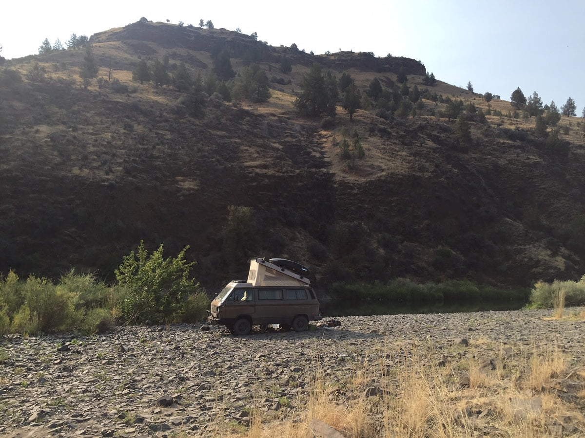 A brown camper van with pop-top tent parked on a rocky riverbank