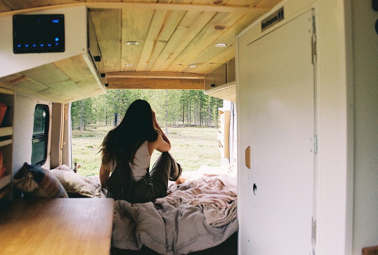 Cozy interior of an RV rental with a person relaxing on the bed, enjoying a serene forest view.