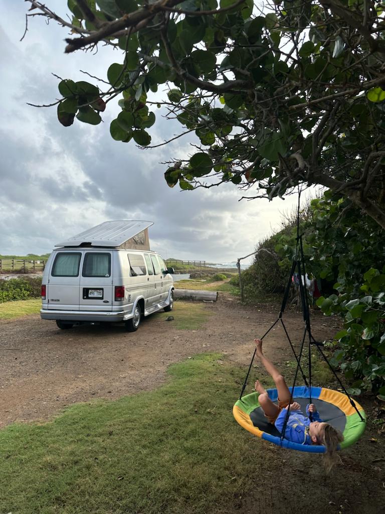 A silver RV rental with a pop-top is parked by the coast. A child relaxes in a colorful saucer tree swing nearby.