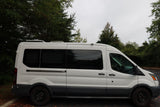 White Ford Transit-style campervan, ideal for RV rental, parked under cloudy skies among trees.