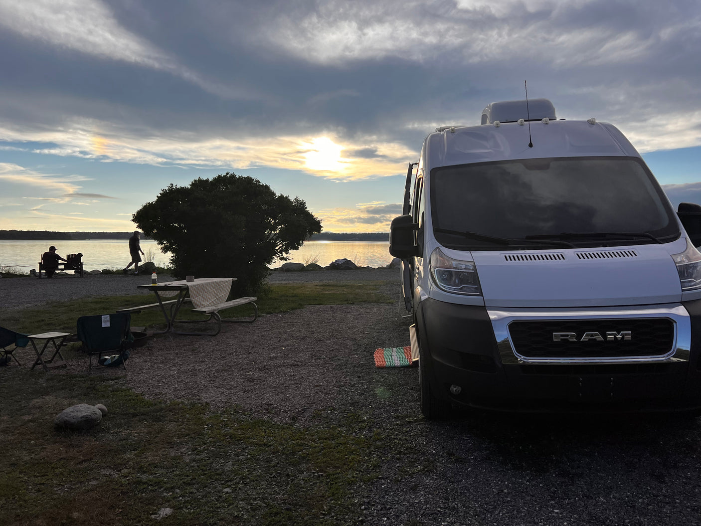 Enjoy an RV rental: a white camper van at a scenic waterfront campsite with a picnic table, chairs, and sunset glow.