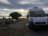 Enjoy an RV rental: a white camper van at a scenic waterfront campsite with a picnic table, chairs, and sunset glow.