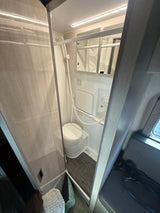 An RV rental's compact wet bath, featuring a toilet, shower, mirror, and bright LED lighting, visible from the living area.