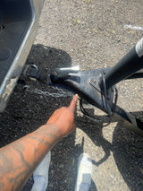 A person points to the RV rental's trailer hitch, showing the secure connection and safety chains on a gravel surface.