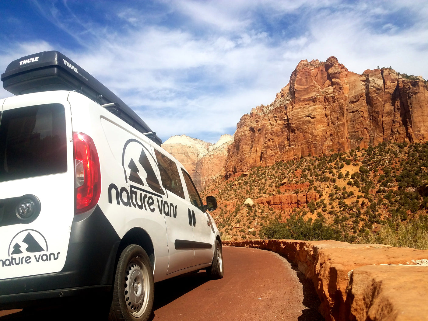 Nature Vans RV rental with Thule box on scenic red rock road. Ideal for park adventures under blue skies.