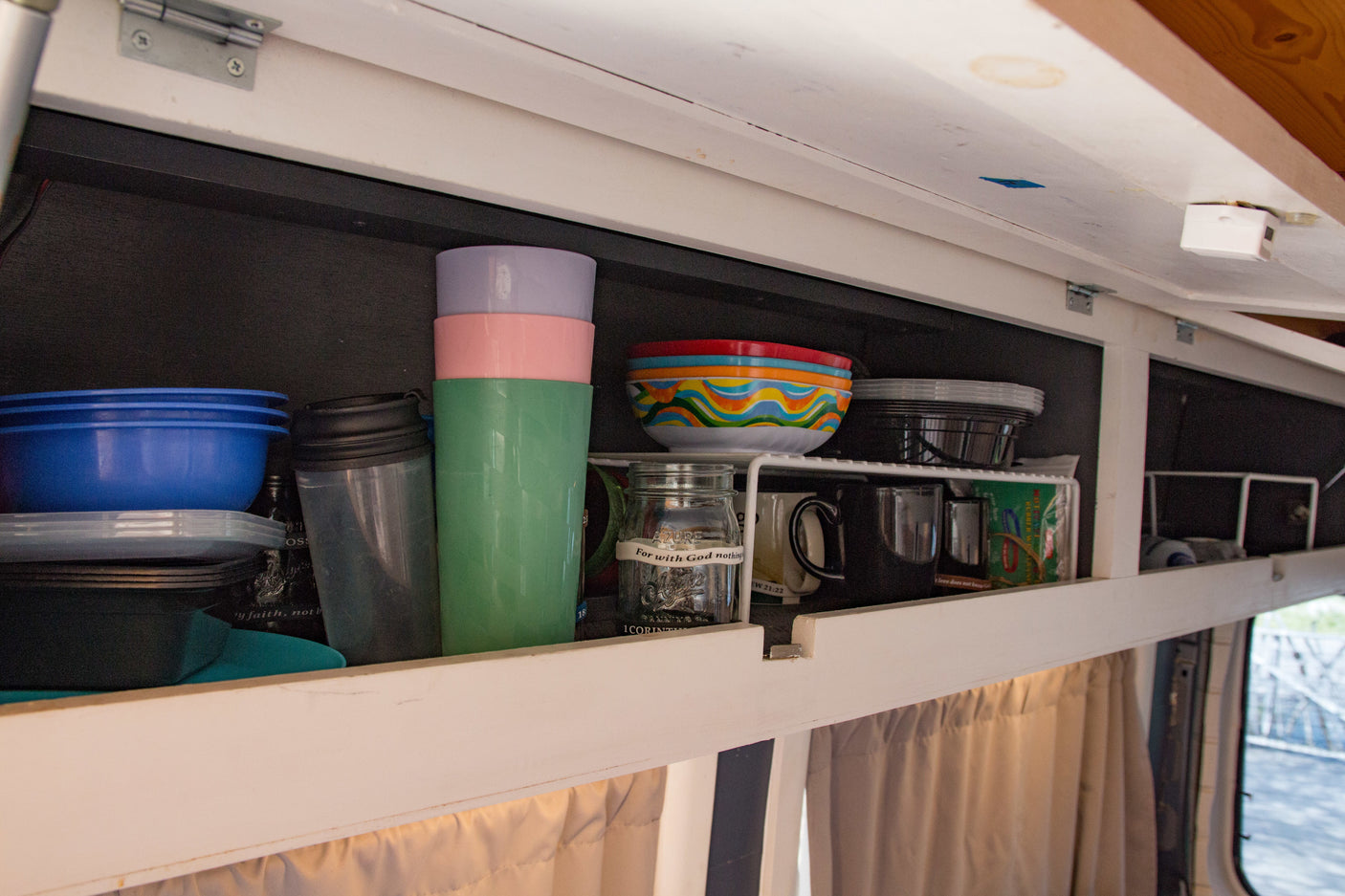 Inside an RV rental, an open overhead cabinet displays neatly organized kitchenware like colorful bowls, cups, and mugs.
