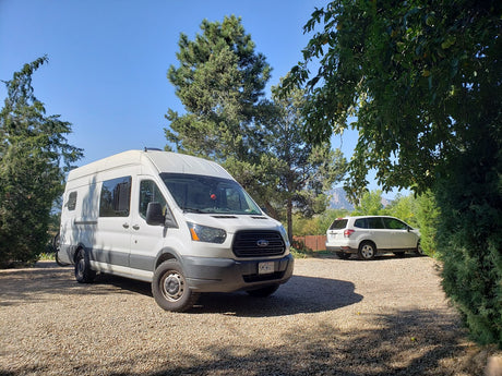 A white Ford Transit camper van RV rental is parked on a sunny gravel driveway with another SUV, trees, and mountains.