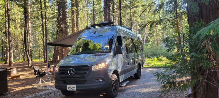 Perfect RV rental for a forest getaway. Gray camper van under an awning at a sunny campsite amidst tall trees.